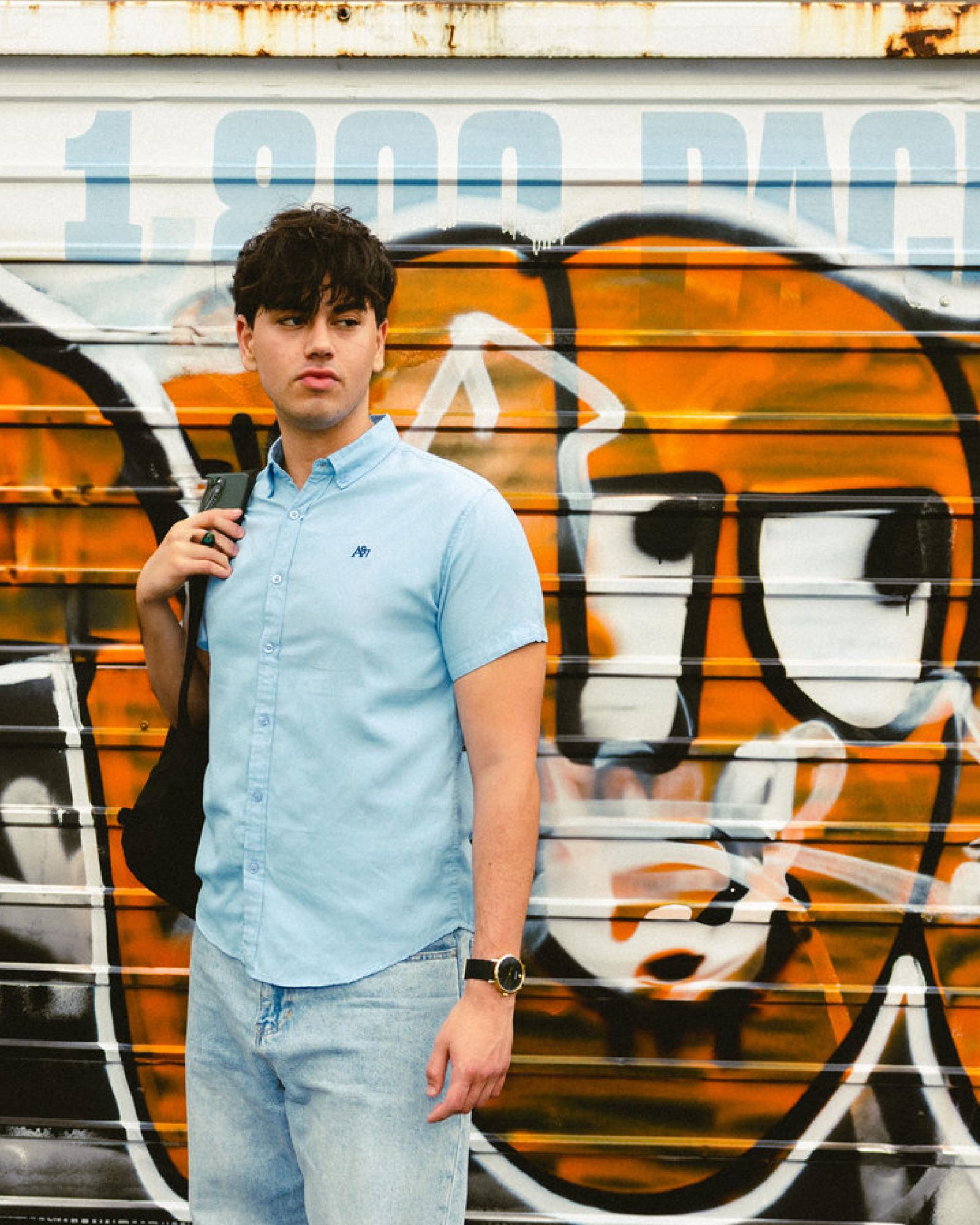Young man in a light blue shirt stands in front of a colorful graffiti wall, holding a backpack strap.
