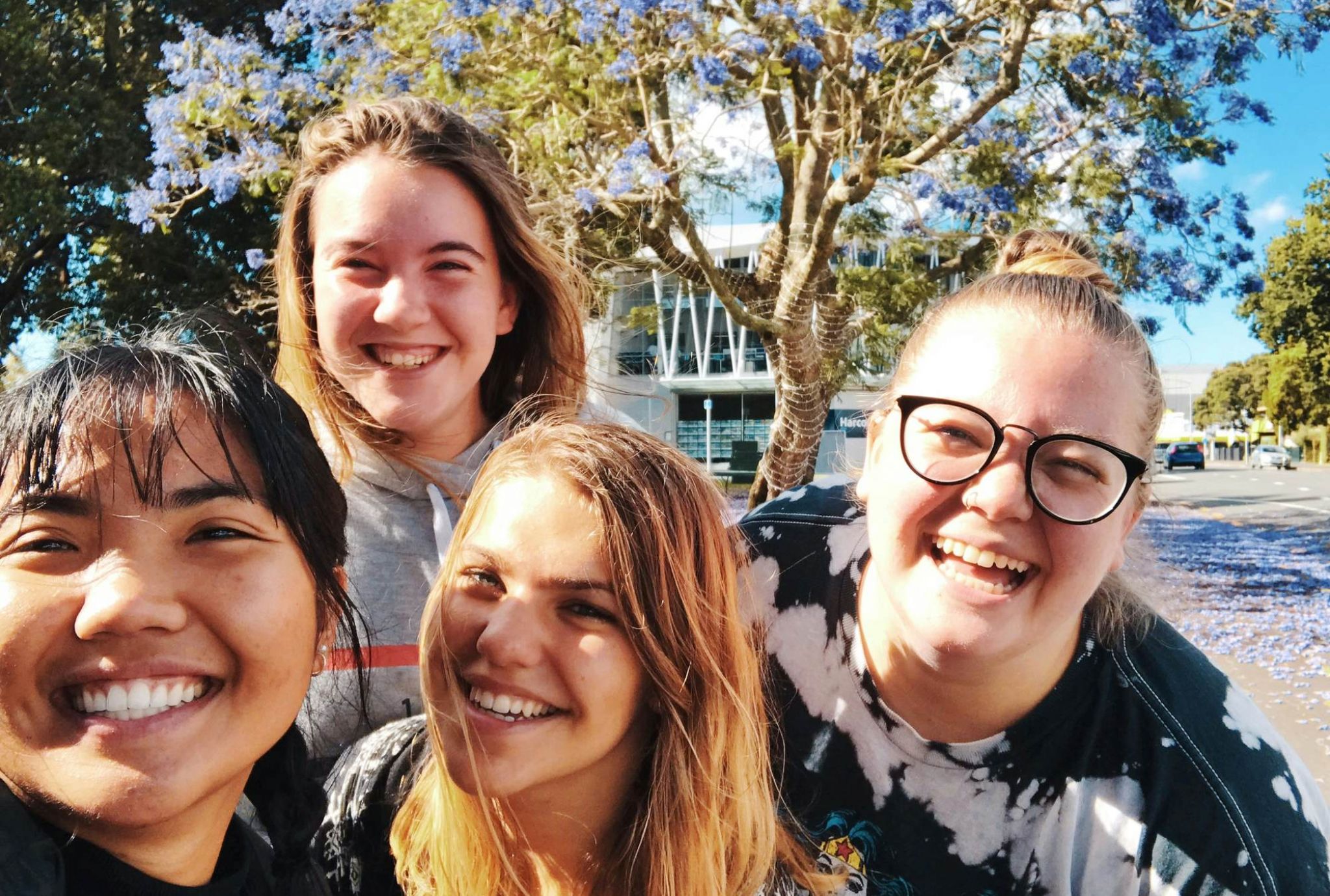 Four young women smiling outdoors under a blooming tree with purple flowers on a sunny day.