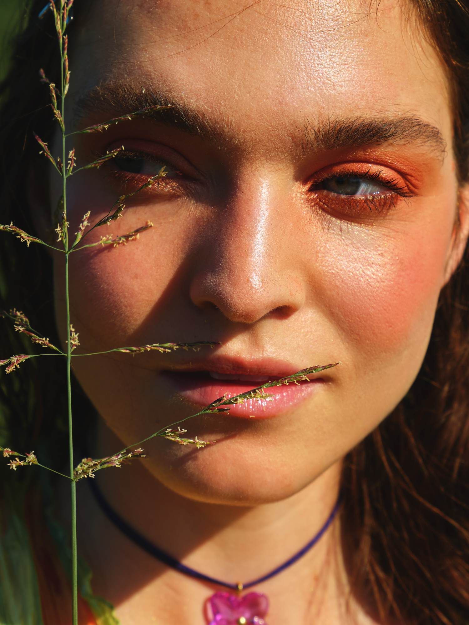 A woman with bold orange eye makeup and a flower necklace, with grass stems in front of her face.