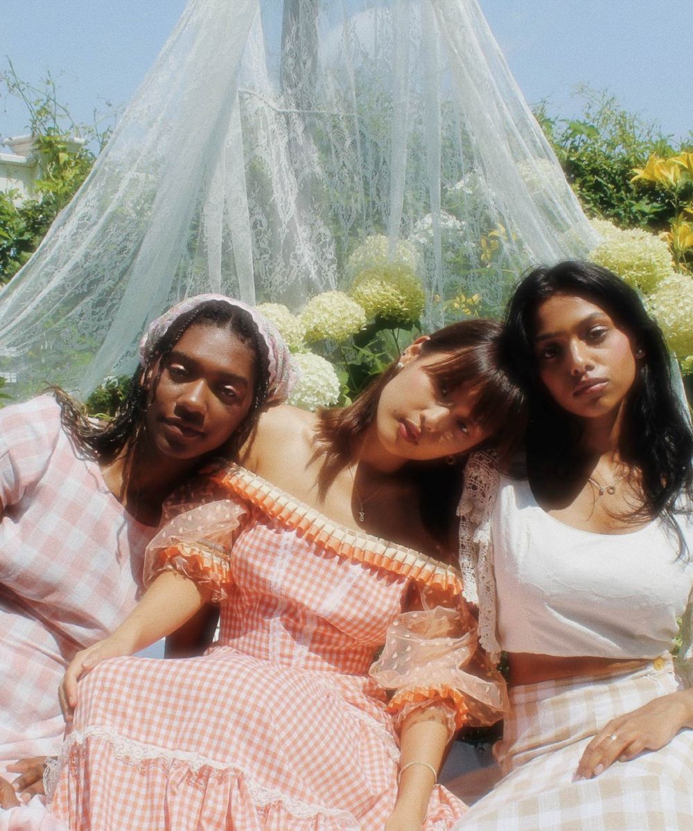 A group of three women sitting together in a sunny garden, dressed in soft gingham and lace outfits, posing in front of flowers and sheer fabric at VERVE Fayetteville.