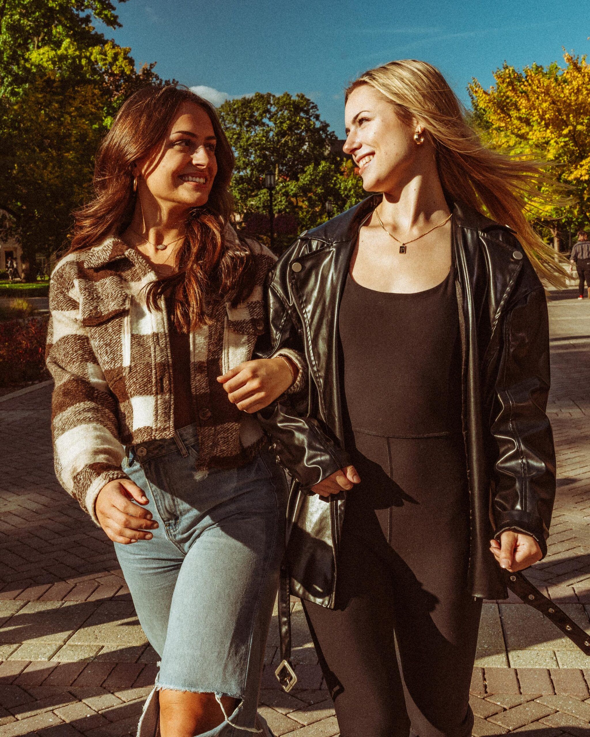Two women, smiling and linking arms, walk outside on a sunny day with trees and blue sky in the background.