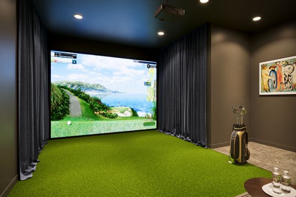 Indoor golf simulator room with a green turf floor, golf clubs, and a large screen displaying a coastal golf course.