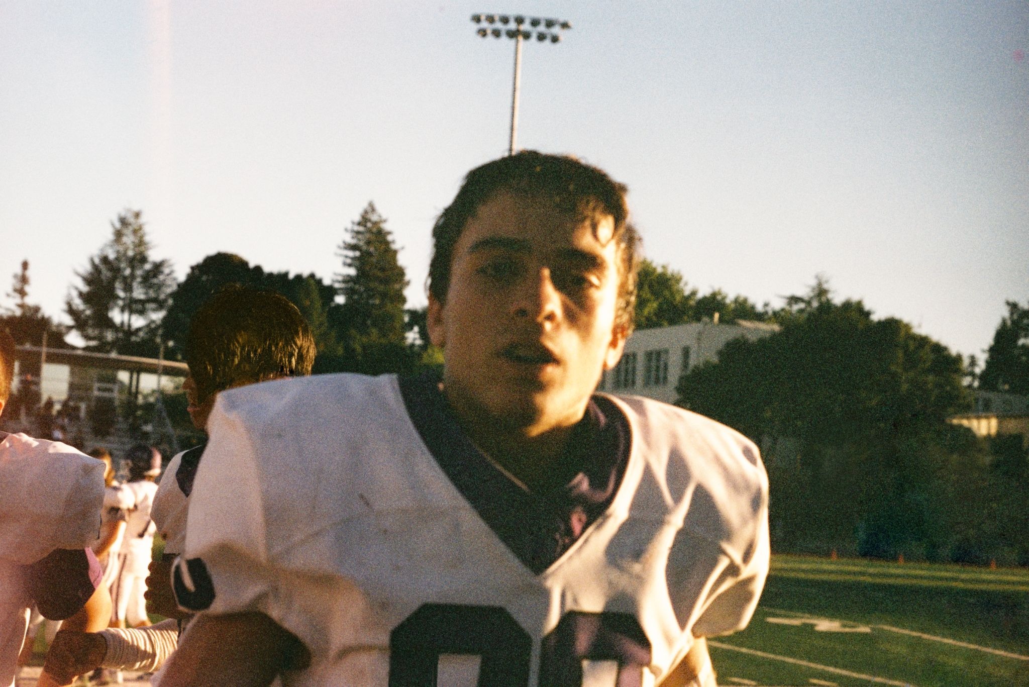A football player in uniform stands on the field, looking toward the camera in the sunlight.